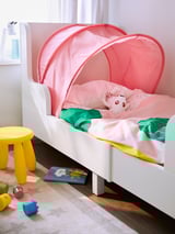 Unha cama infantil cun baldaquino de tenda vermello en forma de cúpula, sabas rosas e un peluche. Un taburete amarelo e xoguetes dispersos están preto.