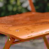 Mesa de aire libre de metal laranxa con gotas de auga, parcialmente dobrada, rodeada de verde.