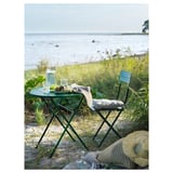 Mesa e cadeira verde ao aire libre colocadas xunto á auga, con bebidas e sombreiro.