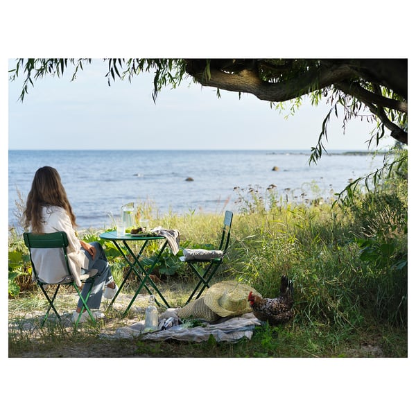 A persoa relaxa na beira do mar con mesa e cadeiras plegables verdes SUNDSÖ, baixo a árbore, con galiña e sombreiro preto.