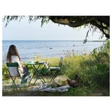 A persoa relaxa na beira do mar con mesa e cadeiras plegables verdes SUNDSÖ, baixo a árbore, con galiña e sombreiro preto.
