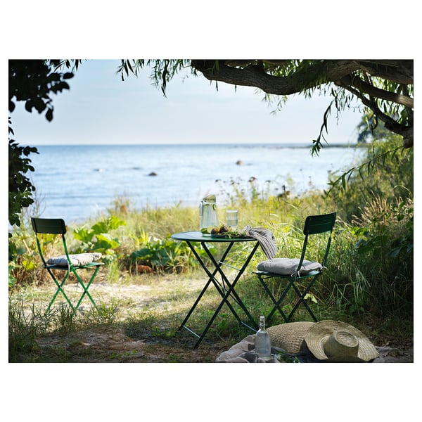 Pequena mesa exterior colocada xunto ao mar, con cadeiras metálicas verdes e mesa con coxíns brancos.