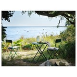 Pequena mesa exterior colocada xunto ao mar, con cadeiras metálicas verdes e mesa con coxíns brancos.