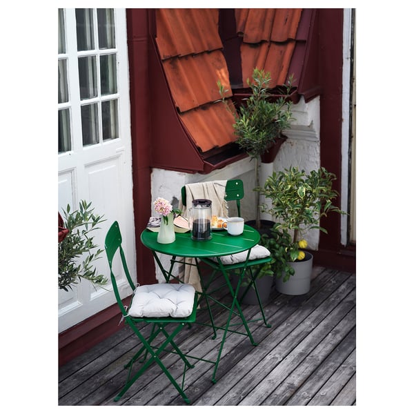 Pequeno patio metálico verde con mesa redonda, dúas cadeiras e coxíns nunha cuberta de madeira xunto a un edificio vermello con plantas.