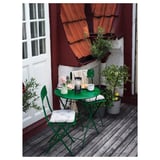 Pequeno patio metálico verde con mesa redonda, dúas cadeiras e coxíns nunha cuberta de madeira xunto a un edificio vermello con plantas.