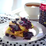 Un prato de waffles rematado con marmelada de blueberry e blueberries frescos, acompañado dunha cunca de café.