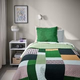 Unha cama cunha cuberta de cama verde TALLSVÄRMARE, xunto a unha pequena mesa cunha lámpada, libros e unha imaxe enriba.