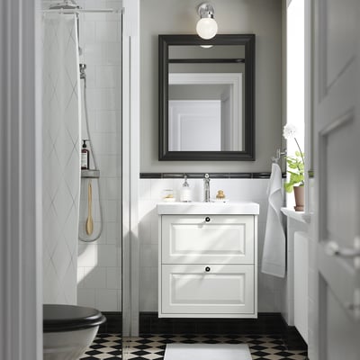 Baño elegante con vanidade branca TÄNNFORSEN, 2 caixóns, lavabo cerámico, espello con marco negro, ducha de vidro, toallas e planta.