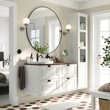 Baño moderno con vaidade branca TÄNNFORSEN, parte superior de madeira, espello redondo, baldosas hexacoadecimalas, plantas e almacenamento.
