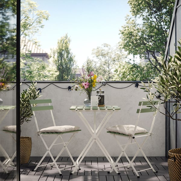 Pequeno balcón exterior con mesa de metal branco e dúas cadeiras, coxíns suaves, vaso con flores e luces de corda.