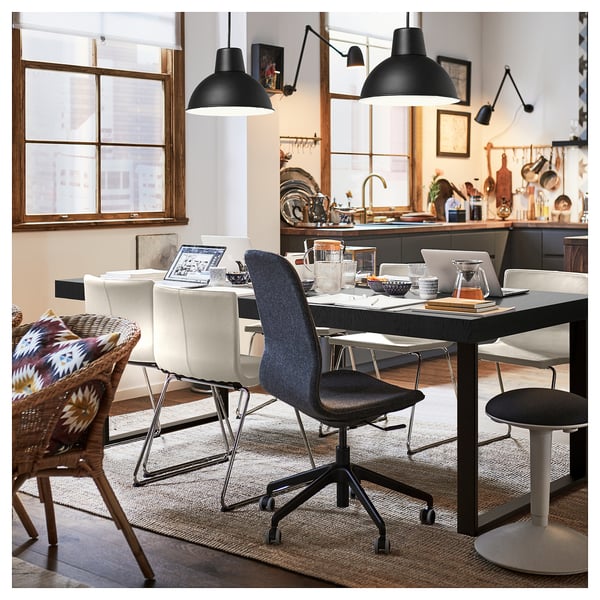 Zona de comedor moderna con revestimento negro TARSELE e mesa extensible metálica, cadeiras brancas, asento azul, lámpadas colgantes negras e cociña ao fondo.