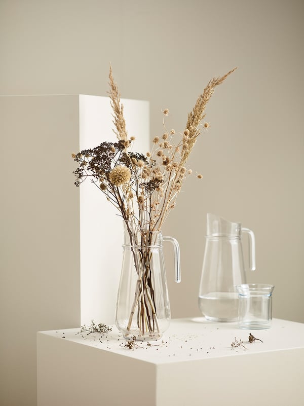 Vaso de vidro transparente con flores secas, dúas xerras de vidro correspondentes, superficie branca, fondo claro.
