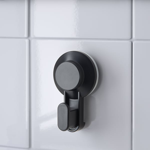 Gancho de ventosa negro montado nunha parede de baldosa branca, ideal para colgar obxectos en baños ou cociñas.