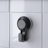 Gancho de ventosa negro montado nunha parede de baldosa branca, ideal para colgar obxectos en baños ou cociñas.
