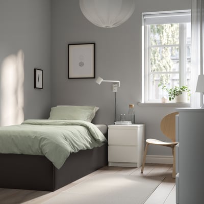 Dormitorio minimalista con cama gris TJÅLLE, roupa de cama verde claro. Mesa de noite branca, cadeira de madeira. Gran fiestra, arte, luz colgante.