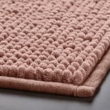 Primeiro plano de alfombra de baño rosa suave e texturizada, mostrando o seu material peluche e de secado rápido.