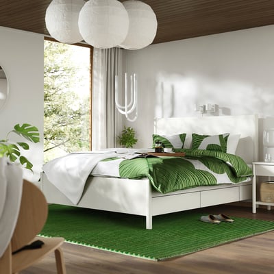Dormitorio con cama branca, roupa verde, luces, alfombra, cadeira, plantas.