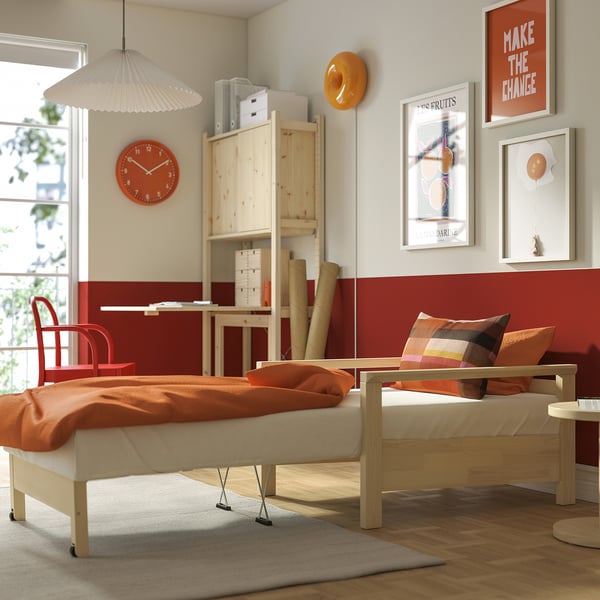 Dormitorio luminoso con cama TORNSBORG dobrada como cadeira de brazos, manta laranxa, reloxo vermello, arte e escritorio de madeira.