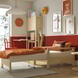 Dormitorio luminoso con cama TORNSBORG dobrada como cadeira de brazos, manta laranxa, reloxo vermello, arte e escritorio de madeira.