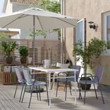 Conxunto de patio moderno con mesa branca TORPARÖ, catro cadeiras azuis correspondentes, gran paraugas beige, plantas e valado de privacidade de madeira.