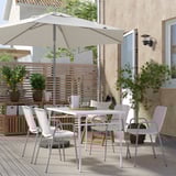 Conxunto de comedor ao aire libre moderno con mesa e cadeiras de metal branco, paraugas beige e plantas na cuberta de madeira.