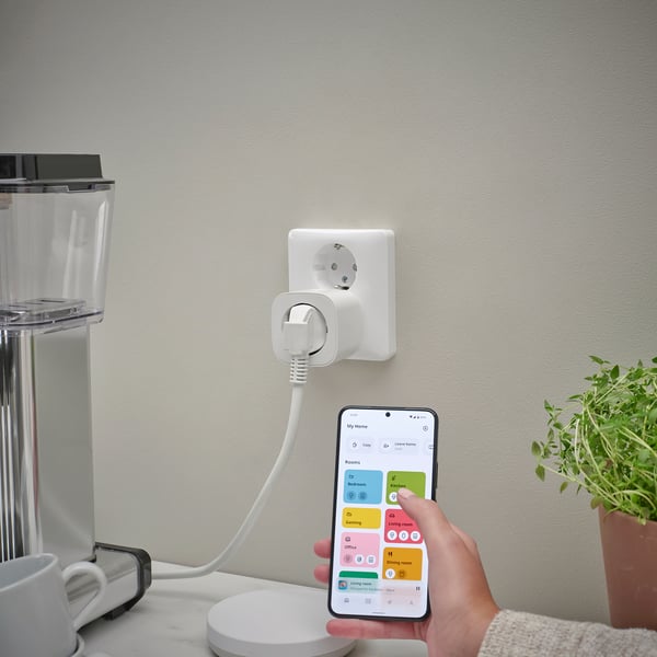 Unha persoa sostén un teléfono intelixente que controla un enchufe conectado a unha cafeteira a través dunha aplicación de fogar intelixente, demostrando a xestión remota do dispositivo.