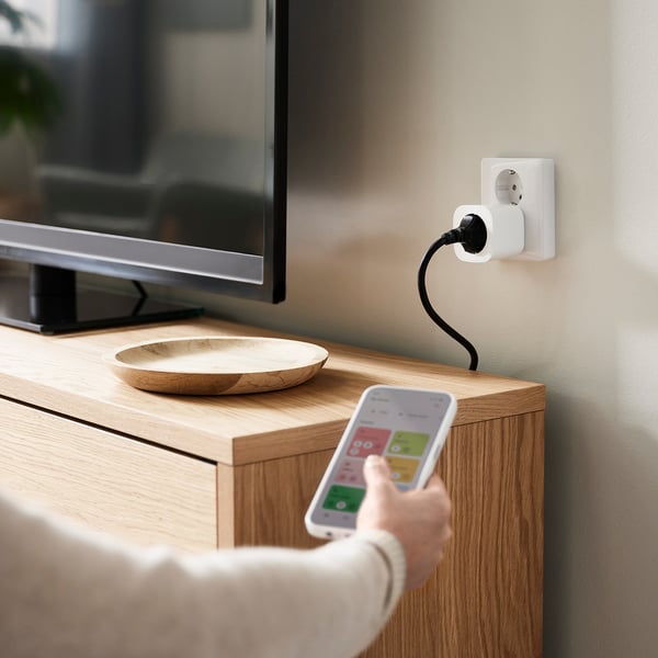 Unha persoa que sostén un teléfono intelixente cunha aplicación domótica, controlando un enchufe intelixente branco conectado a un armario de madeira, cunha cunca e un televisor na parte superior.
