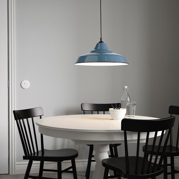 Mesa de comedor branca con cadeiras negras, gran lámpada azul enriba, fondo de parede gris.