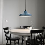Mesa de comedor branca con cadeiras negras, gran lámpada azul enriba, fondo de parede gris.