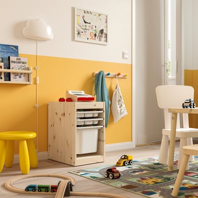 Sala de xogos infantís luminosa con almacenamento TROFAST en madeira natural e branco. Caixas de malla e plástico, un taburete amarelo e xoguetes de madeira.