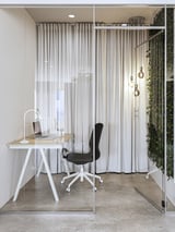 Oficina minimalista moderna cun escritorio TROTTEN beige con pernas en forma de A, unha cadeira negra e iluminación elegante, pechada por vidro e cortinas.