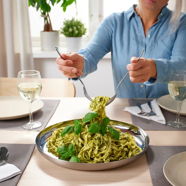 A persoa xira a pasta con garfo e culler ao redor dun prato de servir de aceiro inoxidable redondo, rodeado de vasos de viño branco e pratos.