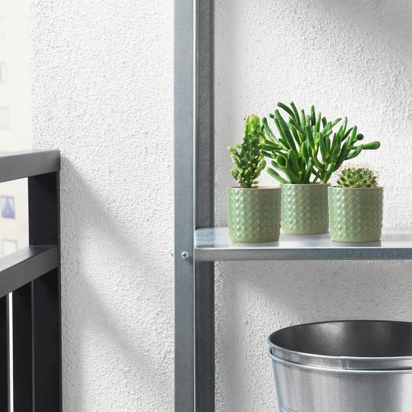 Tres vasos verdes de gres TYSKLÖNN con plantas sentan nun estreito estante metálico xunto a un balde metálico máis grande contra unha parede branca.