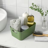 Caixa de almacenamento verde no mostrador, que garda toallas, botellas e cunca. Organiza os elementos de baño de forma ordenada.