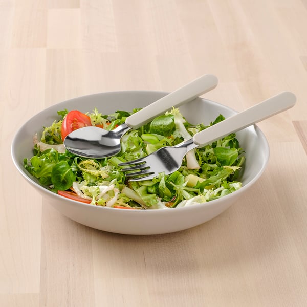 Unha cunca de ensalada fresca con cubertas beige, incluíndo un garfo e culler, colocada sobre unha mesa de madeira.