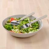 Unha cunca de ensalada fresca con cubertas beige, incluíndo un garfo e culler, colocada sobre unha mesa de madeira.