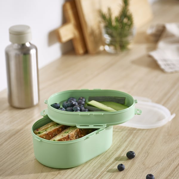 Caixa de xantar ovalada de plástico verde menta UTBJUDA, tamaño similar á botella de auga, dividida en dous niveis para a comida, colocada sobre unha mesa de madeira clara.
