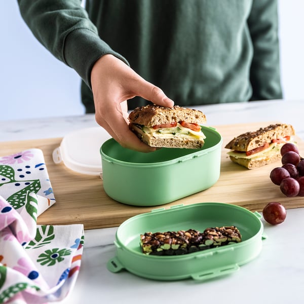 A man coloca o bocadillo nunha caixa de xantar verde de dous niveis, que separa a comida nunha táboa de madeira con uvas e unha servilleta con patróns.