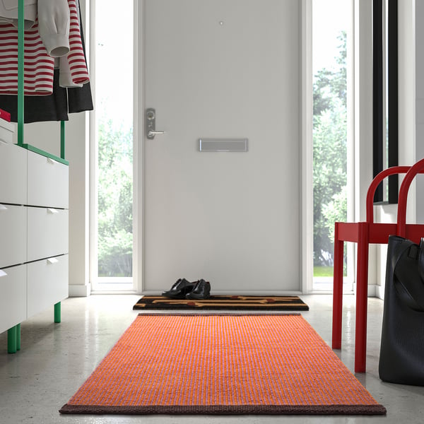 Alfombra VÄGOMRÅDE laranxa e marrón no corredor, preto da porta branca e cadeira vermella.
