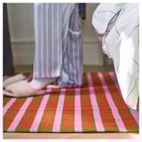 Unha alfombra VÄGSKYLT, con raias rosas e laranxas, feita de algodón. É rectangular, plano e deseñado para aspirar facilmente.