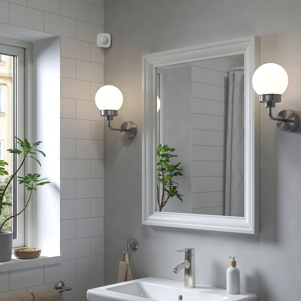 Baño moderno con sensor de movemento VALLHORN sobre lavabo branco, billa de prata, espello e planta. Paredes de azulexos gris claros.