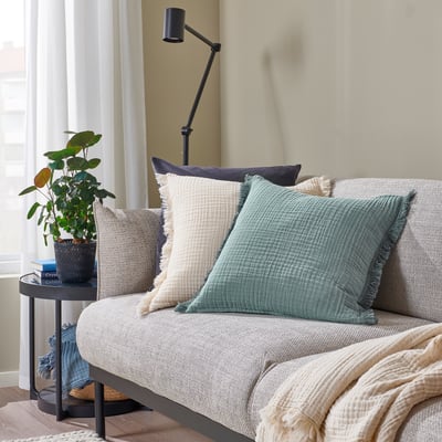 Sala de estar acolledora con sofá beige, coxíns VALLKRASSING, mesa lateral con plantas, luz natural, atmosfera relaxada.