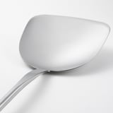 Unha elegante espátula wok de aceiro inoxidable cun mango longo, descansando sobre unha superficie branca sinxela, mostrando o seu deseño pulido e ben elaborado.
