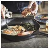 Unha persoa condimentando peixe nunha tixola de ferro fundido negra cun vaso de cervexa ao seu lado.