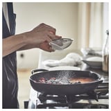 Unha persoa está a cociñar nunha tixola de ferro fundido negra nunha estufa, cribando azucre en po sobre os alimentos.
