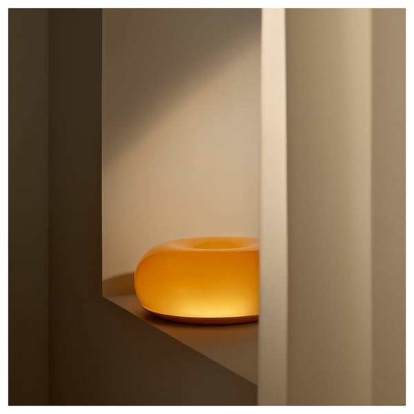 Unha lámpada de mesa de vidro laranxa moderna chamada varmblukt. A súa forma de donuts emite un brillo cálido. A lámpada está situada nun estante minimalista.