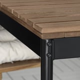 Primeiro plano dunha mesa exterior moderna con tapa de madeira natural e marco de metal negro. Parafusos visibles e construción robusta.