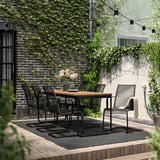 Moderna configuración de comedor ao aire libre con mesa e cadeiras VÄSMAN de metal negro, rodeada de verde e luces de corda.