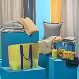 Exhibición de bolsas vaxelbruk, almofadas e lanzamentos en soportes azuis e naturais. Bolsa amarela clara con correas azuis na parte dianteira, máis grande que outros elementos.