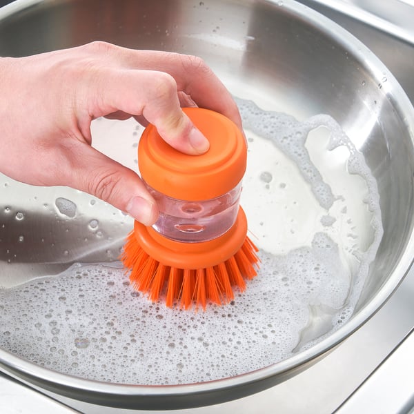 A persoa usa cepillo con mango laranxa e sedas para limpar no lavabo xabón.